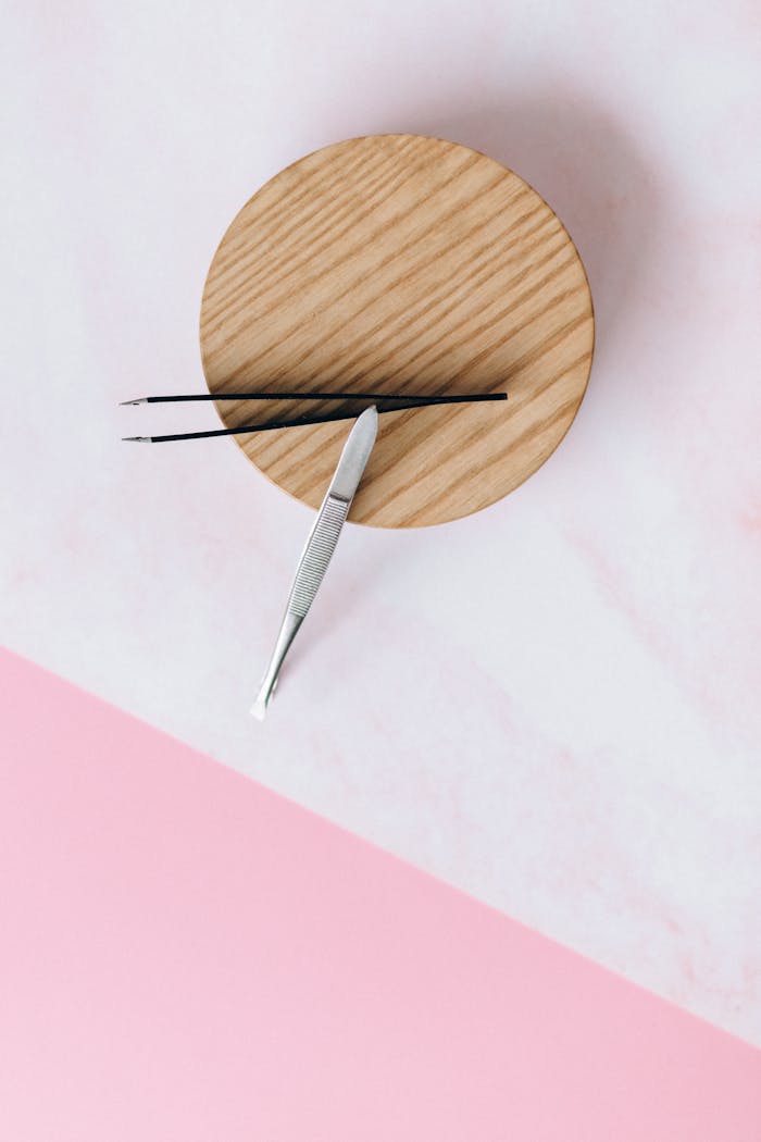 A high-angle shot of tweezers placed on a wooden coaster against a pastel pink background.