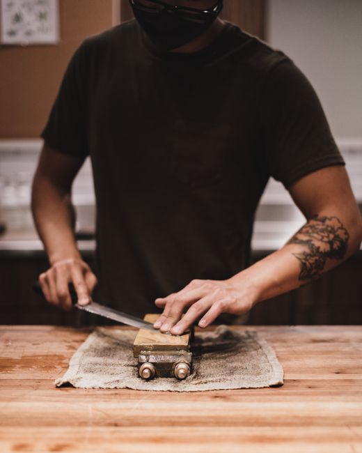 A tattooed man sharpens a knife on a whetstone, honing his skills in a cozy workshop setting.